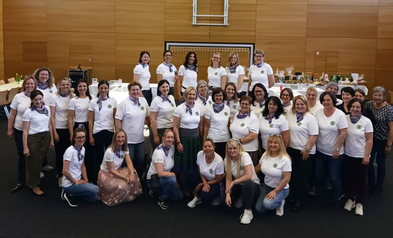 Eine Gruppe von Frauen in weißen Shirts und Schals posiert für ein Foto, einige sitzend und einige stehend in einem Raum mit einer Holzwand.