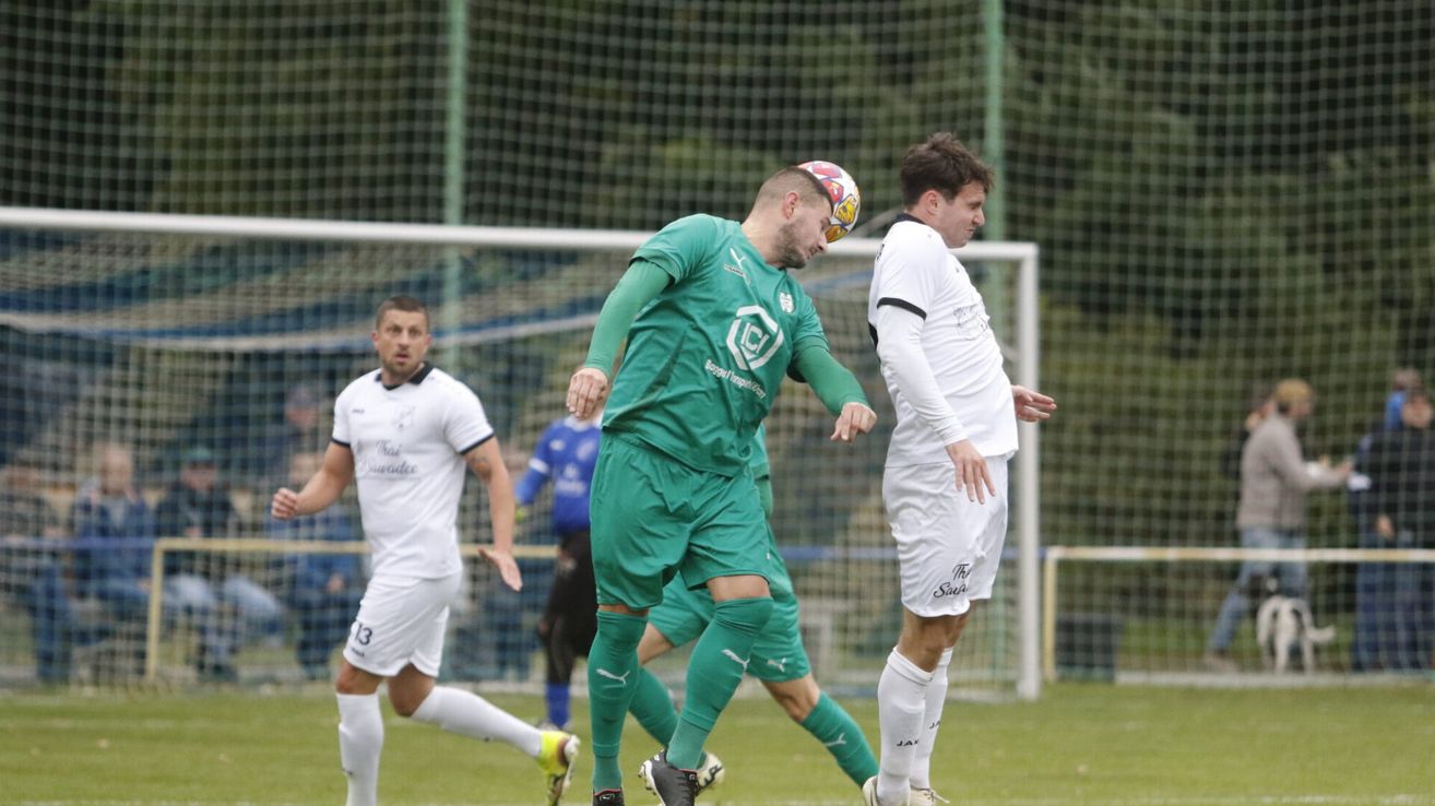Ein Fußballspieler in Grün rennt auf das Tor zu, während ein anderer in Weiß aufmerksam zuschaut. Im Hintergrund laufen zwei weitere Spieler auf einem grünen Feld.