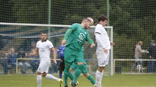Ein Fußballspieler in Grün rennt auf das Tor zu, während ein anderer in Weiß aufmerksam zuschaut. Im Hintergrund laufen zwei weitere Spieler auf einem grünen Feld.