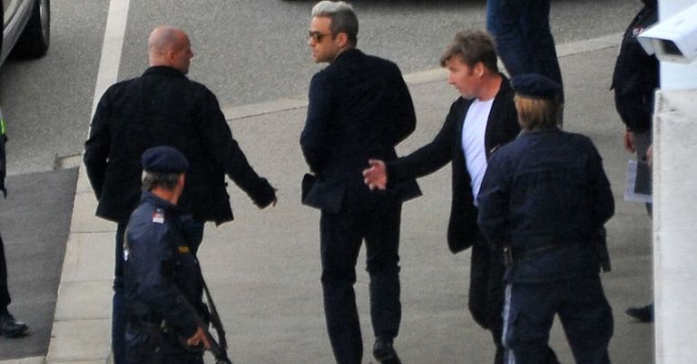 Vier Männer in dunklen Anzügen gehen auf einer Straße, die von Polizisten beobachtet wird. Ein Mann mit Sonnenbrille schüttelt die Hand eines anderen.