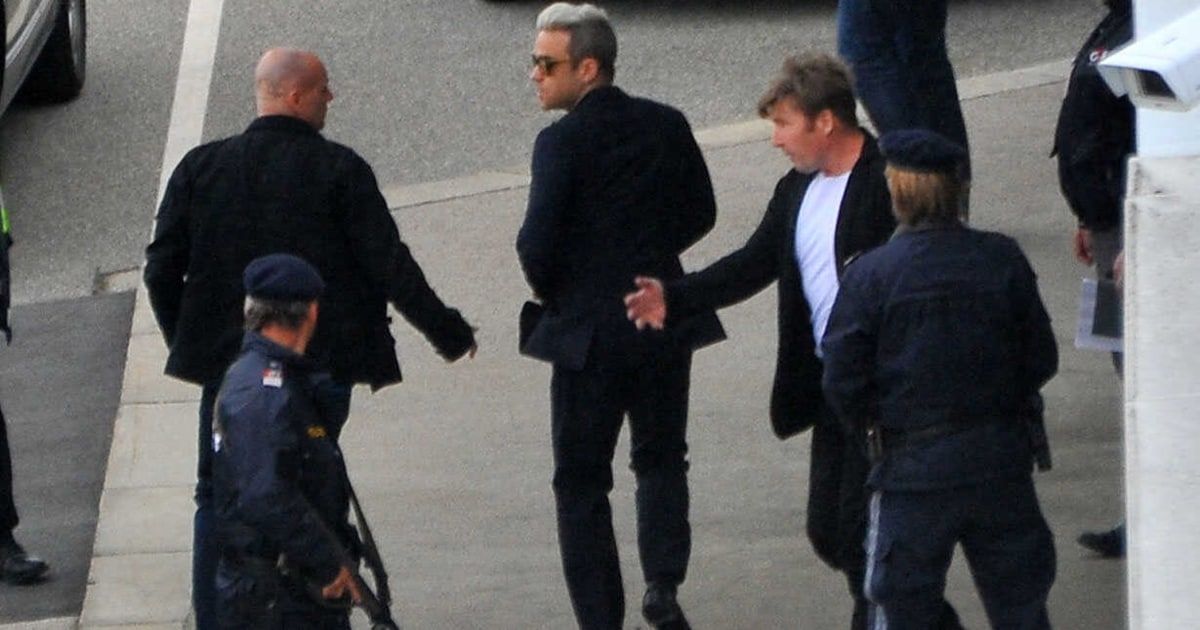 Vier Männer in dunklen Anzügen gehen auf einer Straße, die von Polizisten beobachtet wird. Ein Mann mit Sonnenbrille schüttelt die Hand eines anderen.