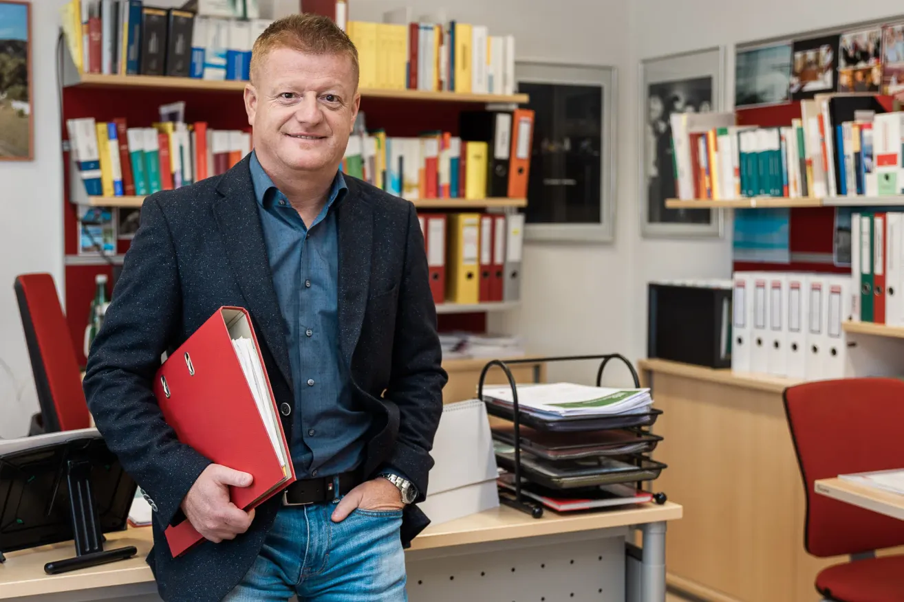 Ein Mann in einem Anzug steht in einem Büro, hält eine rote Mappe in der Hand, lächelt und steht vor einem Bücherregal, das mit Büchern gefüllt ist.