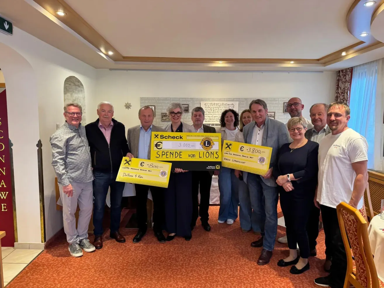 Eine Gruppe von Menschen hält Schecks in einem Raum hoch. Die Schecks stammen vom Lions Club und sind für Beträge von 3.000 und 3.800 Euro ausgestellt. Die Gruppe scheint zu feiern.