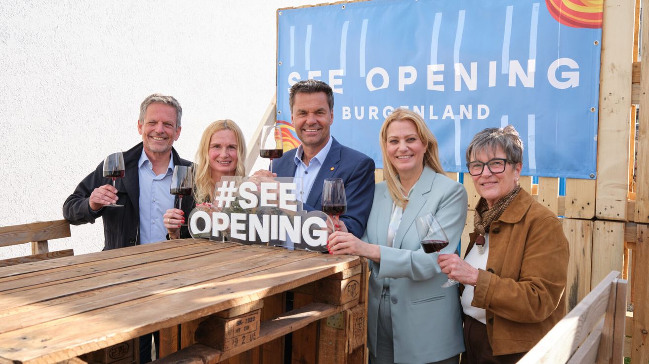 Fünf Personen mit Weingläsern posieren hinter einem Holztresen. Ein Schild dahinter zeigt #SEE OPENING.