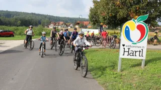 Eine Gruppe von Radfahrern fährt auf einem Pfad, umgeben von Bäumen und einem Willkommensschild. Helme und Sonnenbrillen sind getragen. Dahinter ein Grasbereich und Häuser.