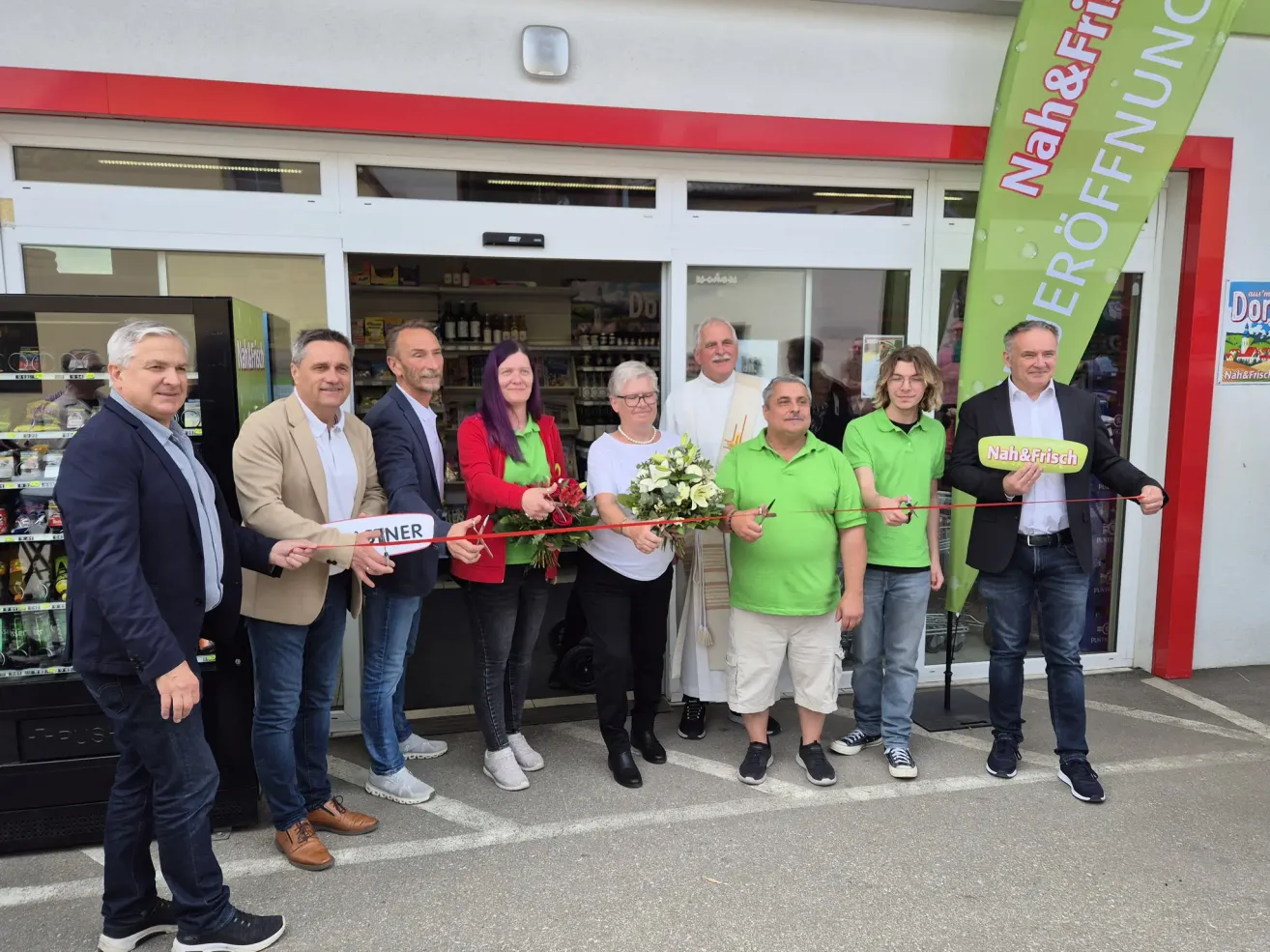 Mehrere Erwachsene, in unterschiedlicher Kleidung, stehen vor einem Geschäft und halten ein Band, wahrscheinlich für eine Ladeneröffnung. Im Hintergrund ist eine Fahne zu sehen.