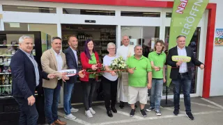 Mehrere Erwachsene, in unterschiedlicher Kleidung, stehen vor einem Geschäft und halten ein Band, wahrscheinlich für eine Ladeneröffnung. Im Hintergrund ist eine Fahne zu sehen.