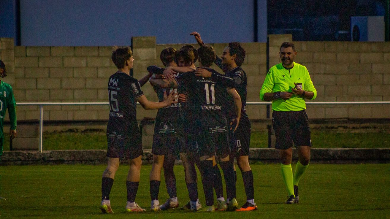 Eine Gruppe von Fußballspielern in schwarzen Trikots feiert auf dem Feld bei Nacht. Der Schiedsrichter steht rechts.
