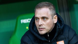 Ein Mann mit kurzen Haaren, der eine schwarze Jacke mit Kapuze und einen orangefarbenen Aufkleber auf der rechten Seite trägt. Er sieht mit ernstem Gesicht nach vorne. Er steht vor einer grünen Wand mit dem Wort 'FEED' darauf.