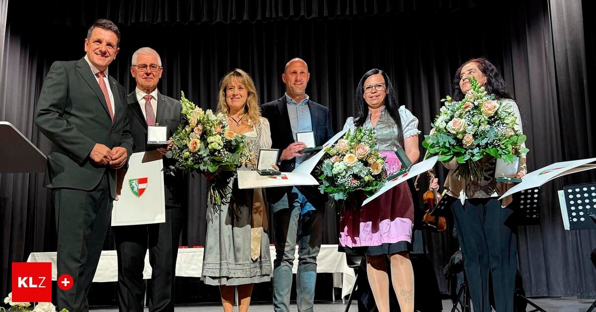 Fünf Personen auf der Bühne halten Blumen und Auszeichnungen und lächeln für ein Foto. Zwei Frauen in traditionellen Kleidern halten Blumensträuße, während Männer Zertifikate halten. Die Bühne hat einen schwarzen Vorhang als Hintergrund.