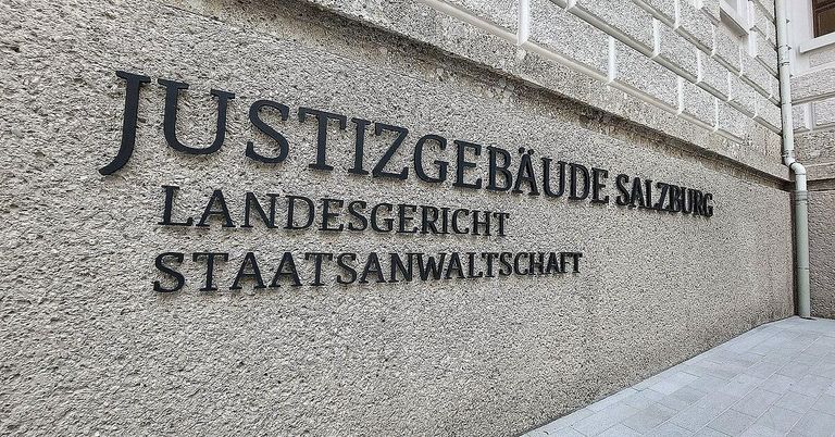 Ein Betongebäude mit dem Text 'JUSTIZGEBÄUDE LANDESGERICHT STAATSANWALTSCHAT'. Ein weißer Handlauf ist an der Wand befestigt.