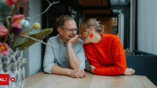 Ein Mann und eine Frau sitzen zusammen an einem Holztisch, lächeln und lachen. Die Frau trägt einen orangefarbenen Pullover, und der Mann trägt eine Brille. Hinter ihnen ist eine Holzwand und eine Pflanze zu sehen.