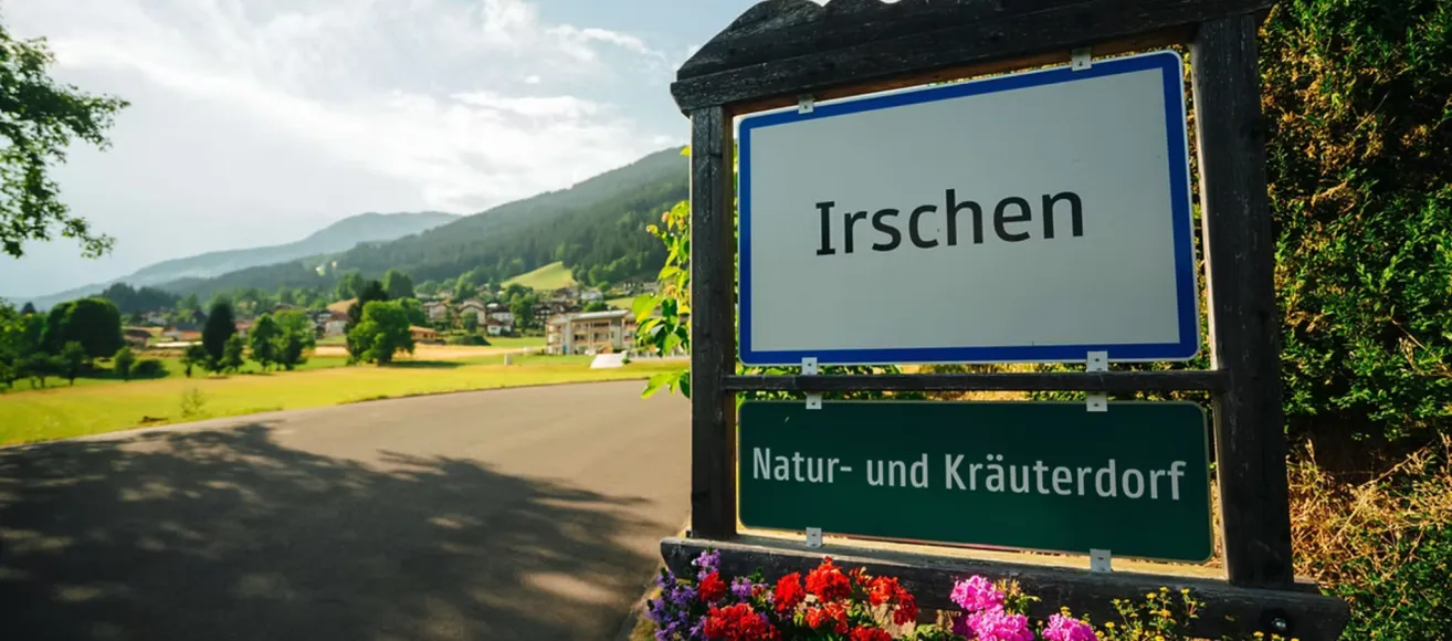 Ein Schild mit der Aufschrift Irschen steht hoch, darunter ein Schild für Natur und Kräuter. In der Ferne sind Berge und Gebäude zu sehen, zusammen mit grünem Gras.