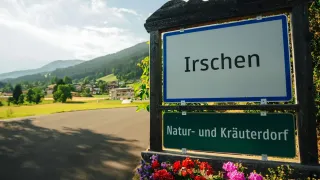 Ein Schild mit der Aufschrift Irschen steht hoch, darunter ein Schild für Natur und Kräuter. In der Ferne sind Berge und Gebäude zu sehen, zusammen mit grünem Gras.