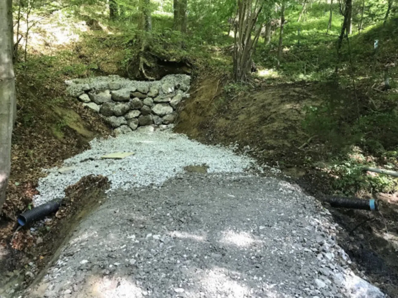 Ein Steinwall wird entlang eines Hangs in einem bewaldeten Gebiet gebaut, mit Kies auf dem Boden.