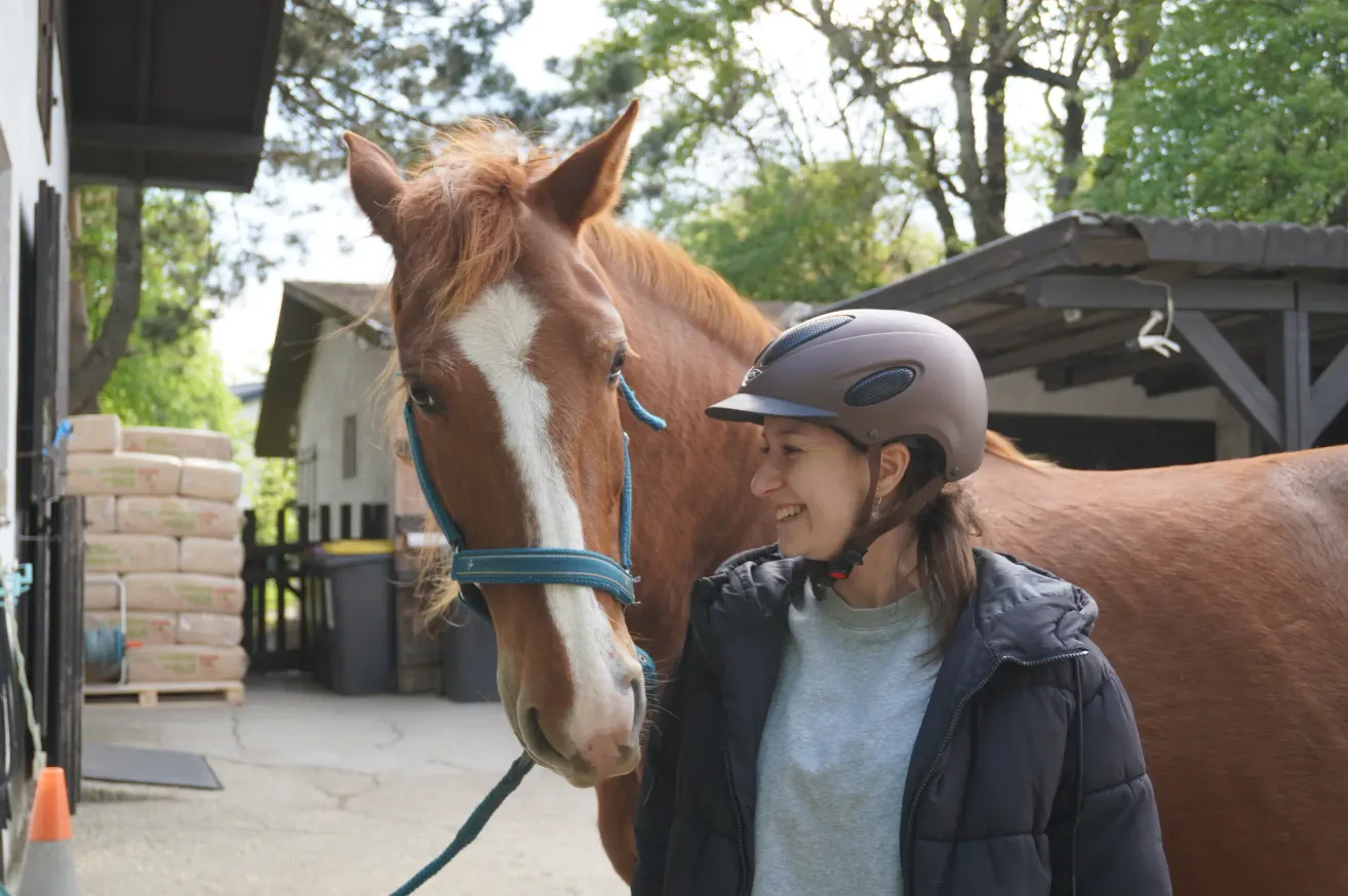 Eine lächelnde Frau mit Helm steht neben einem braunen Pferd mit einem weißen Streifen auf dem Gesicht, an einem sonnigen Ort im Freien.