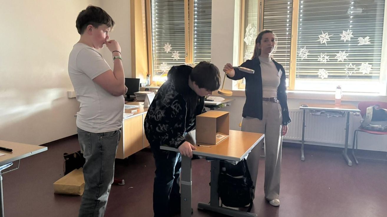 Drei Personen in einem Raum. Eine Person beugt sich über eine Kiste, eine andere hält eine Paddel, und die dritte beobachtet.