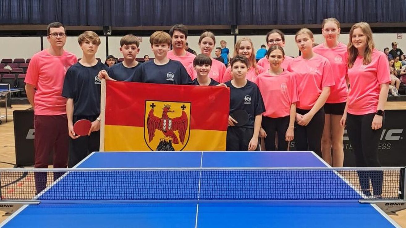 Eine Gruppe junger Leute in passenden Uniformen posiert mit einer Flagge bei einem Tischtennis-Event.