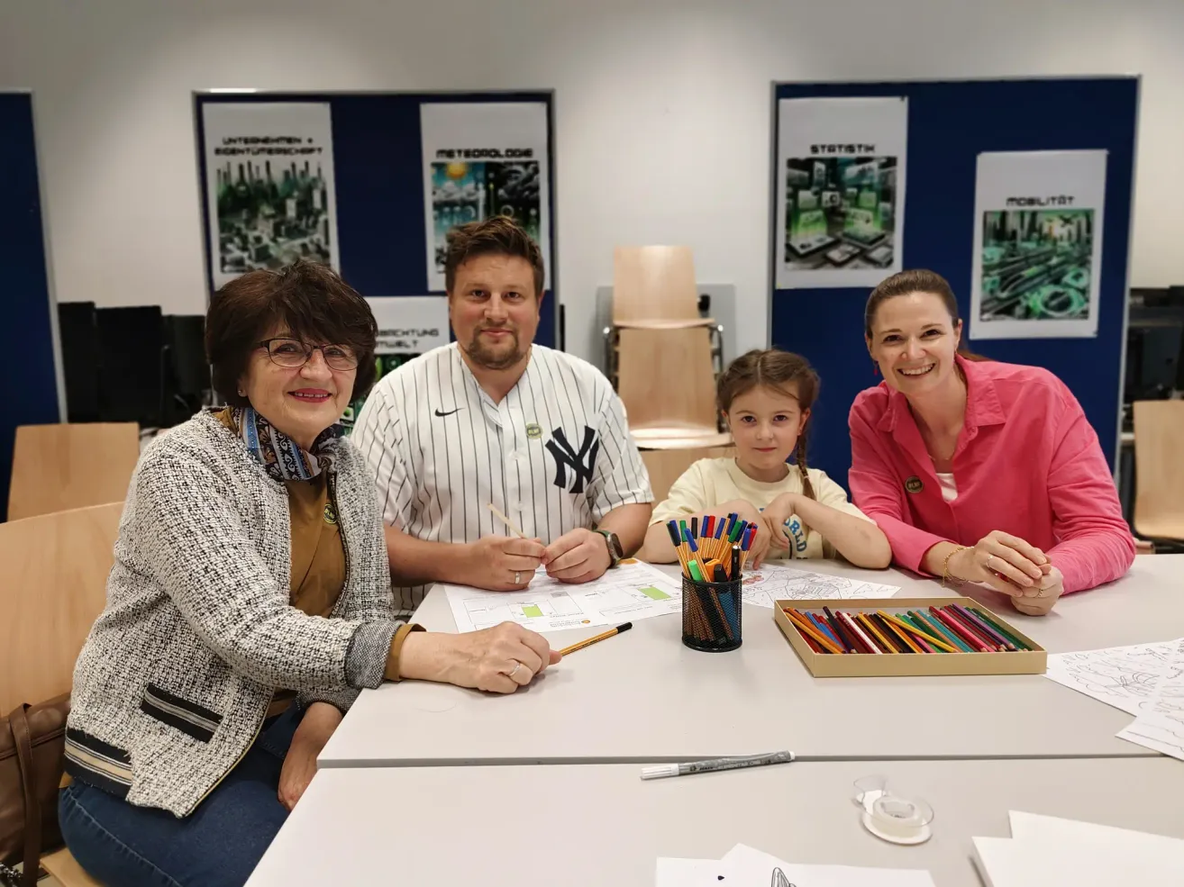 Eine Familie sitzt an einem Tisch. Ein Mann in einem Yankees-Hemd zeichnet. Eine Frau und ein Mädchen lächeln und schauen zu. Papiere, Stifte und eine Schachtel Buntstifte liegen auf dem Tisch.