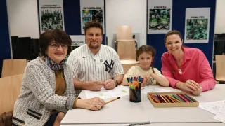 Eine Familie sitzt an einem Tisch. Ein Mann in einem Yankees-Hemd zeichnet. Eine Frau und ein Mädchen lächeln und schauen zu. Papiere, Stifte und eine Schachtel Buntstifte liegen auf dem Tisch.