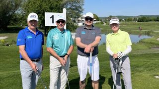 Vier Männer stehen auf einem Golfplatz mit Golfschlägern. Sie lächeln und posieren für ein Foto. Einer trägt ein 'GOLF'-Shirt, ein anderer ein 'GWP'-Shirt, und sie tragen alle Mützen. Hinter ihnen ist ein Golfloch mit einem Nummer-1-Schild.