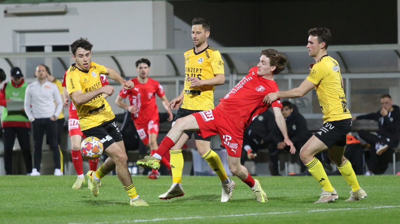 Fußballspieler in roten und gelben Trikots sind in einem dynamischen Spiel verwickelt, wobei ein Spieler in Rot einen Schuss versucht, während andere zuschauen.