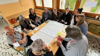 Eine Gruppe von Menschen sitzt im Kreis um einen Tisch und diskutiert. Eine Frau schreibt auf ein Stück Papier.