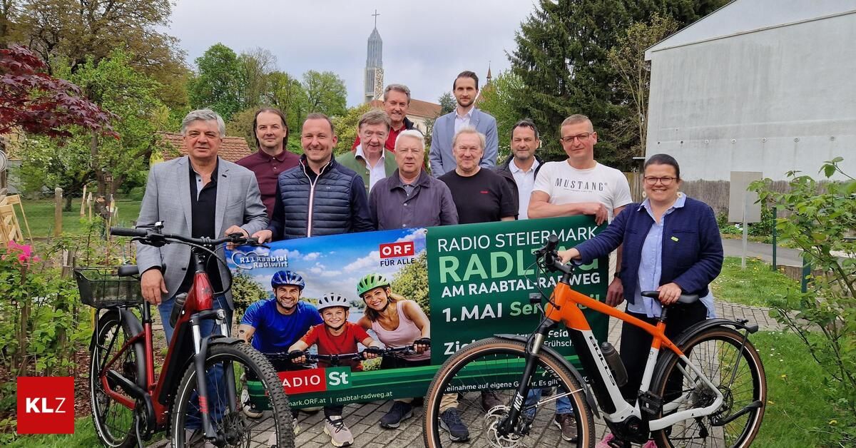 Eine Gruppe von Menschen posiert mit Fahrrädern und einem grünen Banner mit der Aufschrift Radio Steiermark. Sie lächeln und halten das Banner. Bäume und Gebäude sind im Hintergrund zu sehen.