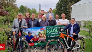Eine Gruppe von Menschen posiert mit Fahrrädern und einem grünen Banner mit der Aufschrift Radio Steiermark. Sie lächeln und halten das Banner. Bäume und Gebäude sind im Hintergrund zu sehen.