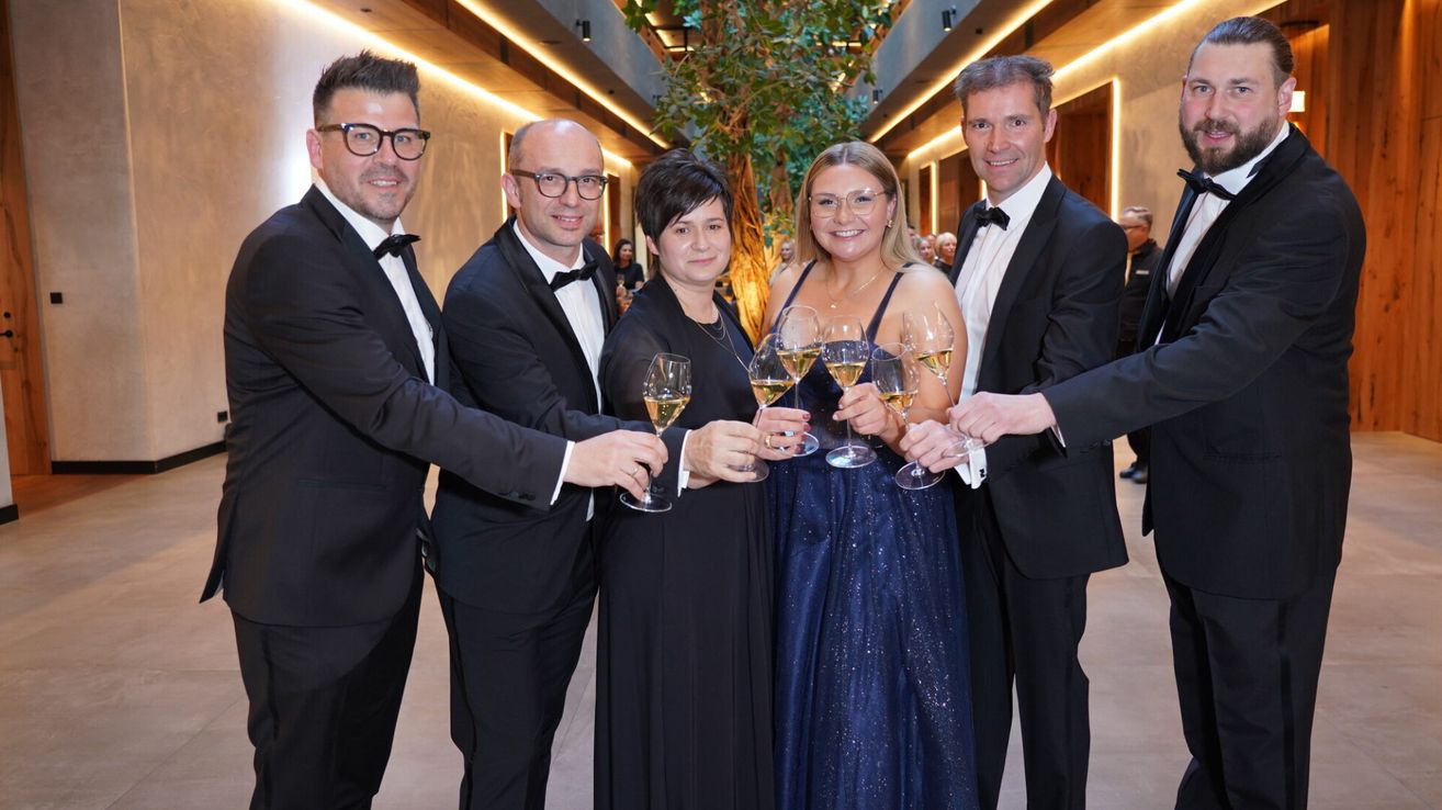 Fünf Menschen in formeller Kleidung posieren für ein Foto und halten Weingläser. Zwei Männer und zwei Frauen lächeln, mit einem großen Baum im Hintergrund.