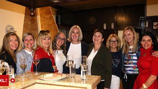 Sieben Frauen stehen um eine Bar herum und lächeln, mit Weingläsern, einem Teller Essen und einer Flasche. Eine Frau trägt einen roten Schal.