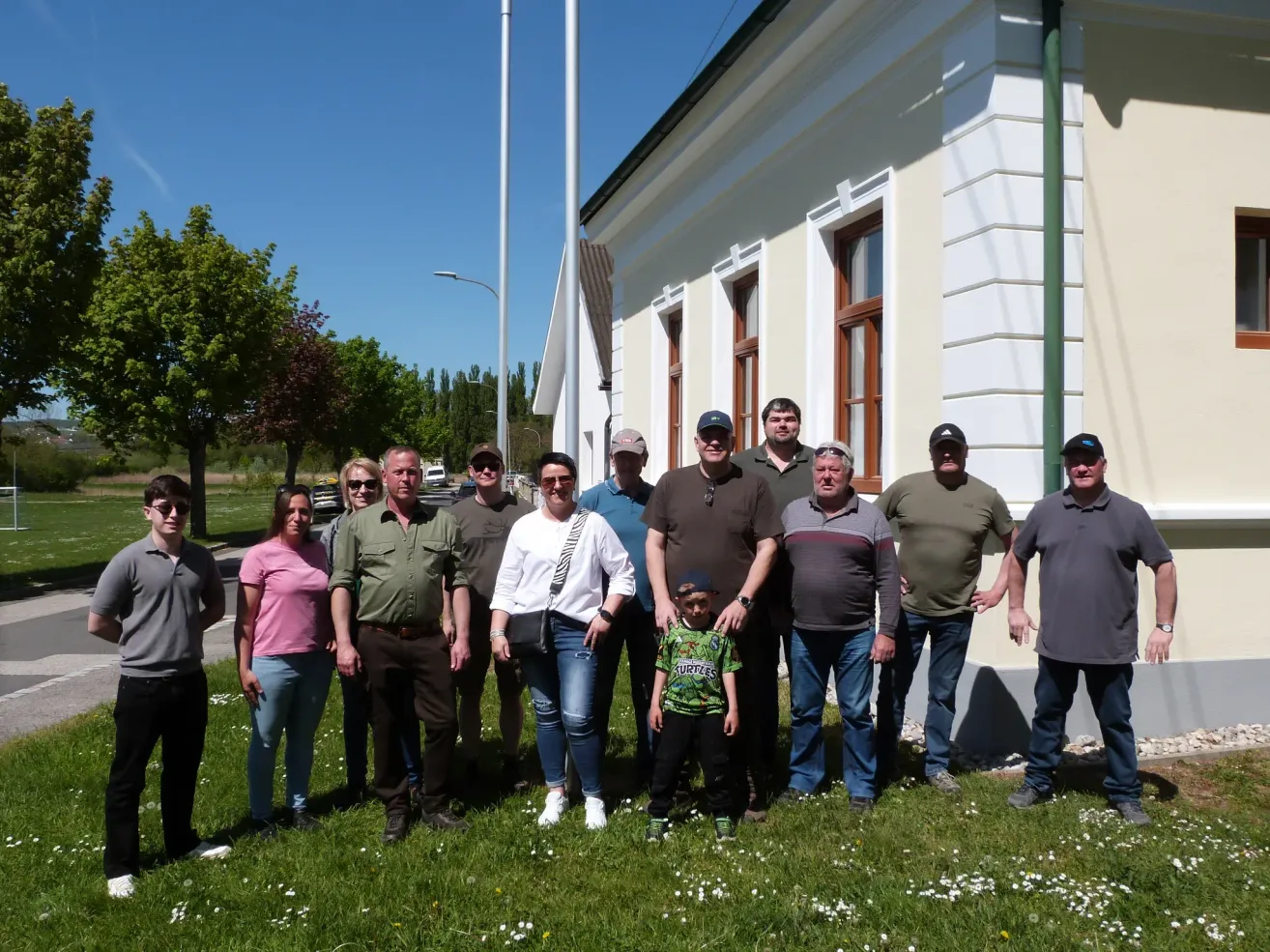 Eine Gruppe von Menschen steht auf einem Rasen vor einem weißen Gebäude. Ein Kind in der Mitte trägt ein grünes Shirt. Alle lächeln. Bäume und ein Pfahl sind im Hintergrund.