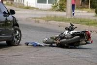 Ein Motorrad liegt auf der Seite auf der Straße, mit einem blauen Tuch in der Nähe. Ein schwarzer Wagen ist am Straßenrand geparkt.