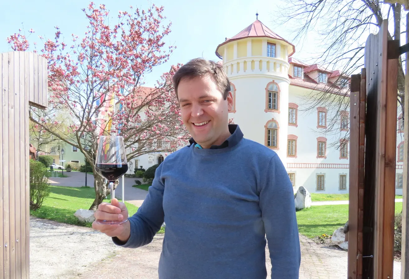 Ein Mann in einem blauen Hemd hält ein Glas Rotwein vor einem historischen Gebäude mit rosa Blumen.
