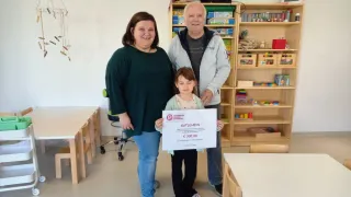 Eine Familie steht in einem Raum mit Regalen voller Spielzeug. Die Frau und der Mann stehen hinter dem Kind, das ein Zertifikat hält. Das Kind hält das Zertifikat mit ernster Miene.