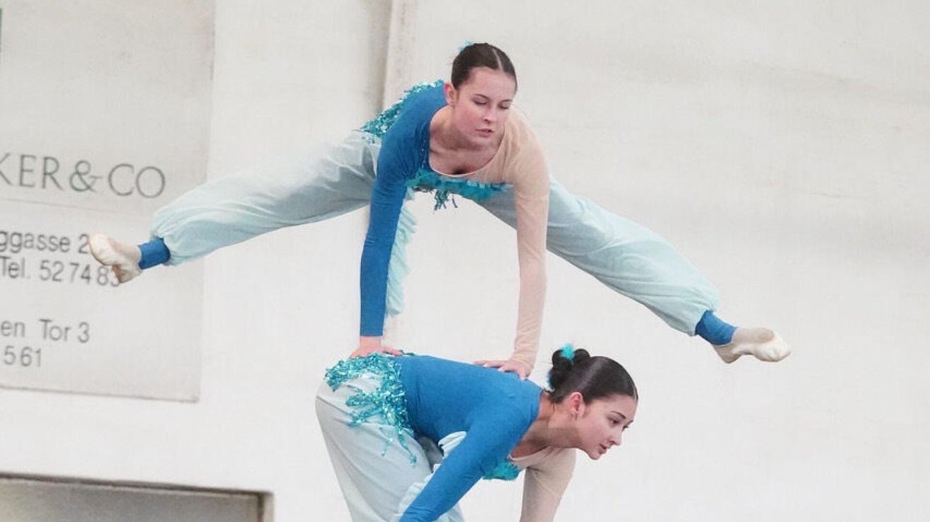 Zwei Frauen führen einen akrobatischen Stunt durch, eine steht auf den Schultern der anderen, beide tragen passende blaue Outfits mit weißen Hosen.