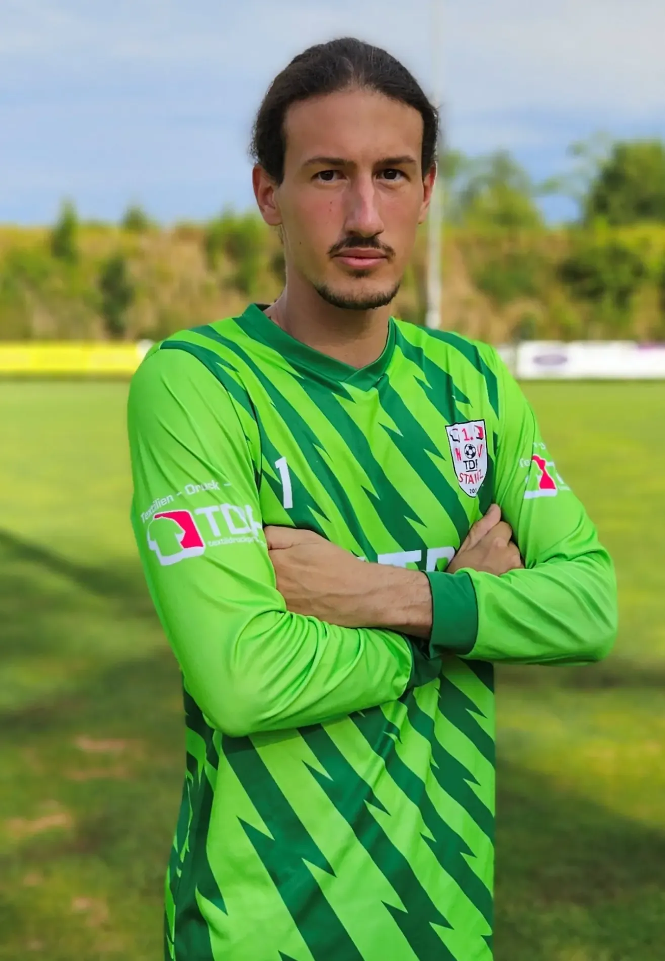 Ein Fußballspieler in einem grünen Trikot mit der Nummer 1 steht auf einem Rasenfeld mit verschränkten Armen.