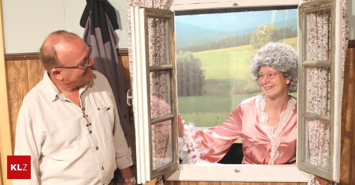 Ein älterer Mann mit Brille und Uhr steht neben einem Fenster mit einem blumigen Vorhang. Eine Frau in einem rosa Nachthemd und Perücke lächelt und schaut aus dem Fenster.