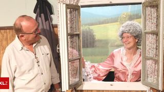 Ein älterer Mann mit Brille und Uhr steht neben einem Fenster mit einem blumigen Vorhang. Eine Frau in einem rosa Nachthemd und Perücke lächelt und schaut aus dem Fenster.