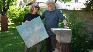 Zwei Personen stehen im Freien und lächeln, wobei eine eine große abstrakte Malerei hält. Sie sind in der Nähe einer Betonskulptur, mit Grünpflanzen und Häusern im Hintergrund.