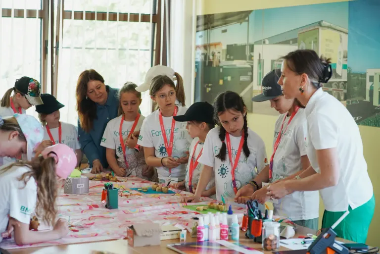 Eine Gruppe von Kindern in weißen Shirts mit Namensbändern versammelt sich um einen Tisch und bastelt und malt. Eine Frau in einem blauen Shirt steht neben ihnen.
