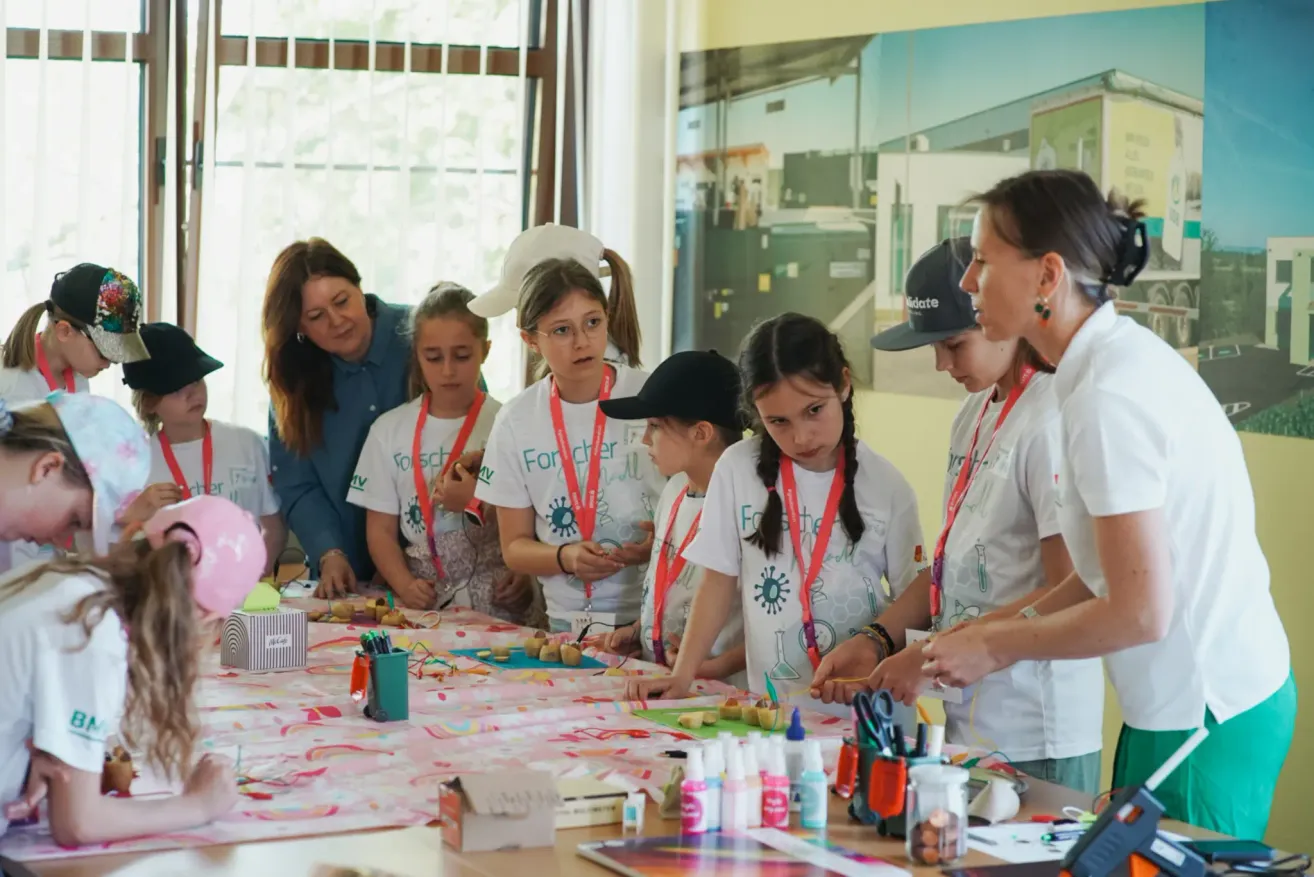 Eine Gruppe von Kindern in weißen Shirts mit Namensbändern versammelt sich um einen Tisch und bastelt und malt. Eine Frau in einem blauen Shirt steht neben ihnen.