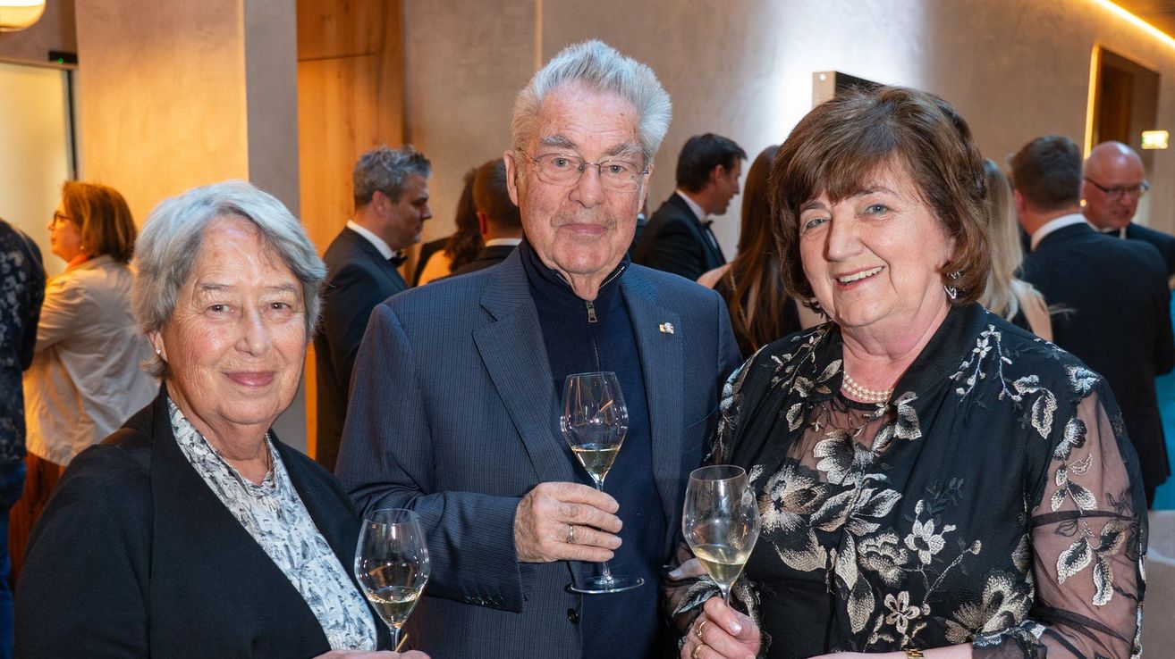 Ein älterer Mann mit Brille und einem blauen Anzug hält ein Weinglas mit Weißwein und steht zwischen zwei lächelnden Frauen. Im Hintergrund stehen mehrere Personen und unterhalten sich.