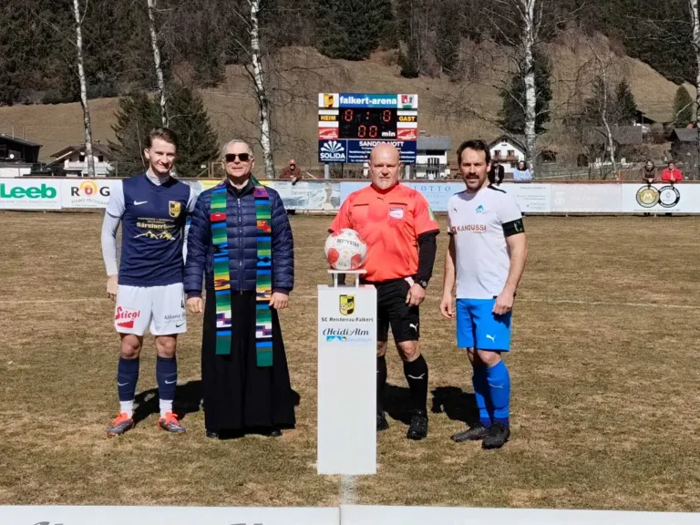 Vier Männer stehen lächelnd auf einem Fußballfeld. Ein Pokal ist zwischen ihnen. Zwei Männer tragen blaue Trikots. Ein Mann trägt ein oranges Shirt. Der vierte Mann trägt eine blaue Robe. Ein Anzeigebrett ist im Hintergrund.