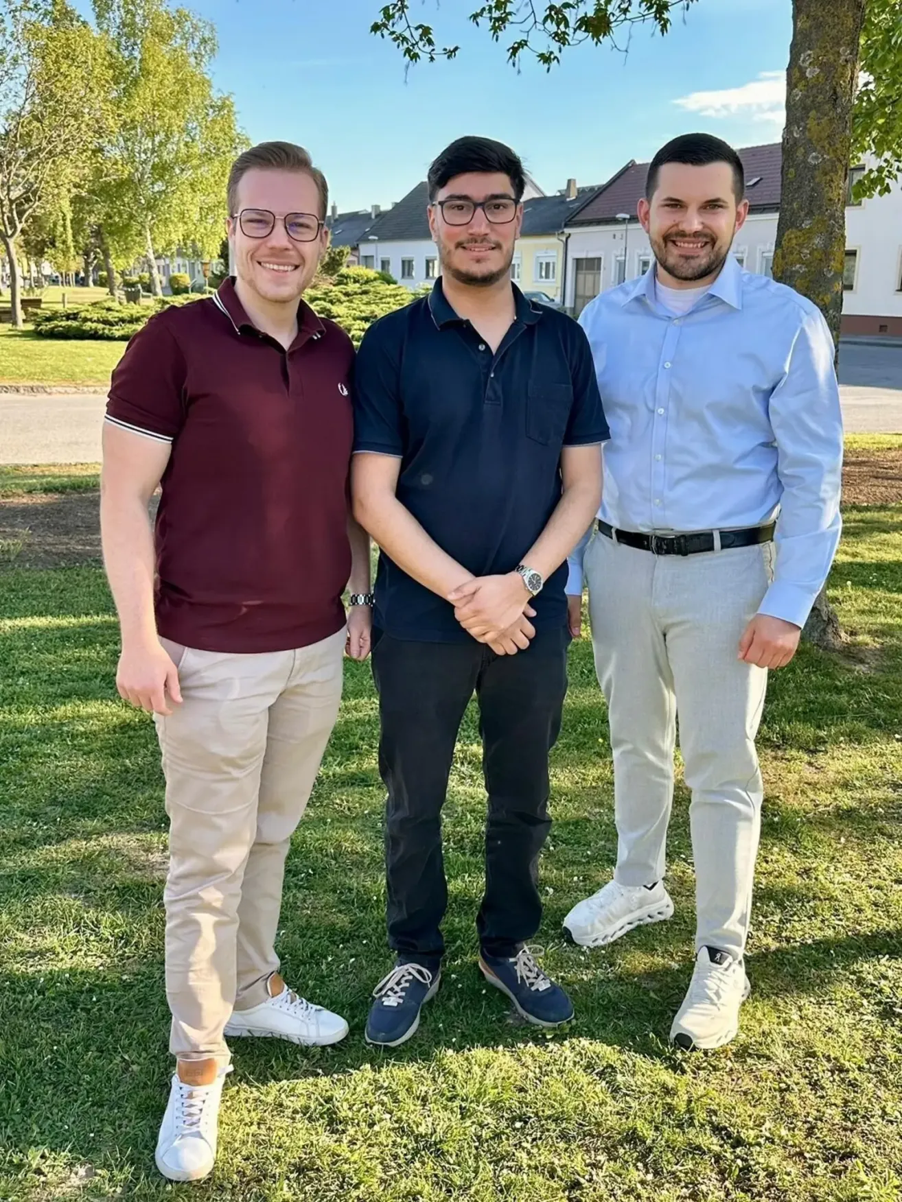 Drei Männer stehen in einem Park. Sie sind lässig gekleidet. Der Mann in der Mitte trägt eine Brille und hat die Hände verschränkt. Der Mann auf der rechten Seite trägt ein blaues Hemd und einen Gürtel. Der Mann auf der linken Seite trägt ein Weinrot-Hemd und eine Brille.