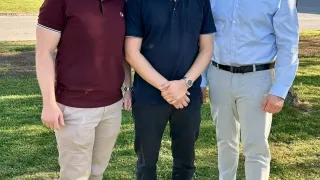 Drei Männer stehen in einem Park. Sie sind lässig gekleidet. Der Mann in der Mitte trägt eine Brille und hat die Hände verschränkt. Der Mann auf der rechten Seite trägt ein blaues Hemd und einen Gürtel. Der Mann auf der linken Seite trägt ein Weinrot-Hemd und eine Brille.