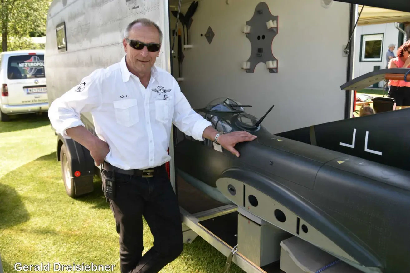 Ein Mann in einem weißen Hemd und Sonnenbrille steht neben einem Jet, seine Hand auf dem Cockpit, mit einem Anhänger hinter ihm.
