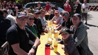 Eine Gruppe von Menschen, einschließlich Erwachsener und Kindern, sitzt um einen Tisch herum und lächelt und posiert für ein Foto. Flaschen und Gläser mit Getränken stehen auf dem Tisch. Im Hintergrund stehen und gehen weitere Menschen.