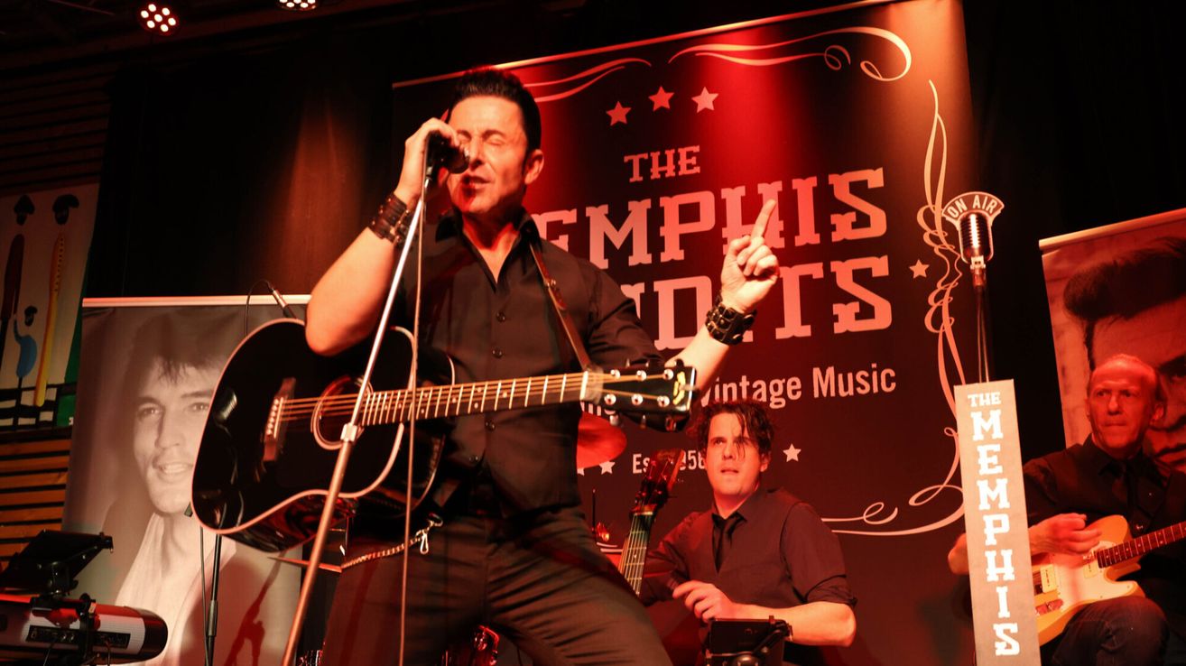 Ein Mann tritt auf der Bühne mit einer Gitarre und einem Mikrofon auf und spielt vor einem Schild, das 'The Memphis Sounds' steht. Ein anderer Mann spielt im Hintergrund ein Instrument.