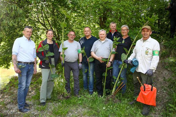 Eine Gruppe von Menschen steht in einem Wald und lächelt für ein Foto. Sie halten Pflanzen und tragen Handschuhe. Eine Frau trägt eine Brille. Sie befinden sich auf einem Pfad in der Nähe eines Flusses.